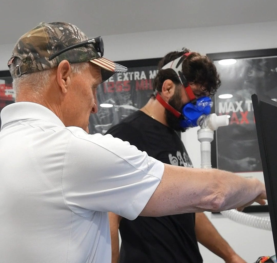 A man in a white polo shirt and camouflage cap points toward another man wearing a blue and red face mask while exercising on a treadmill inside a gym.